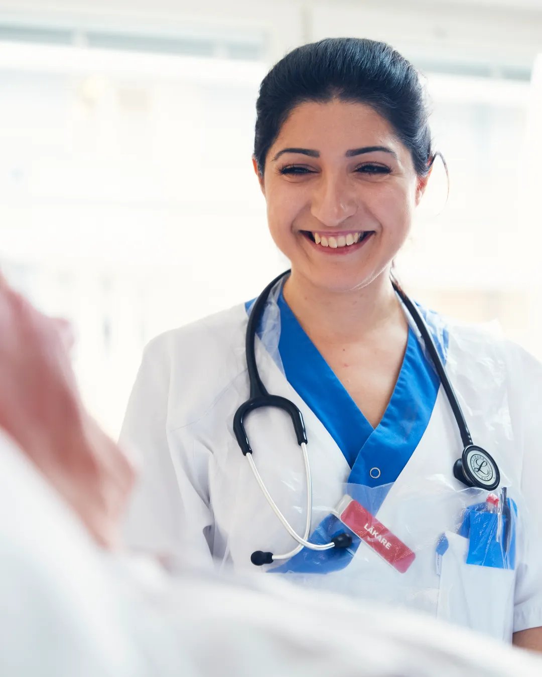 En kvinna i vit rock med stetoskop ler mot patient.