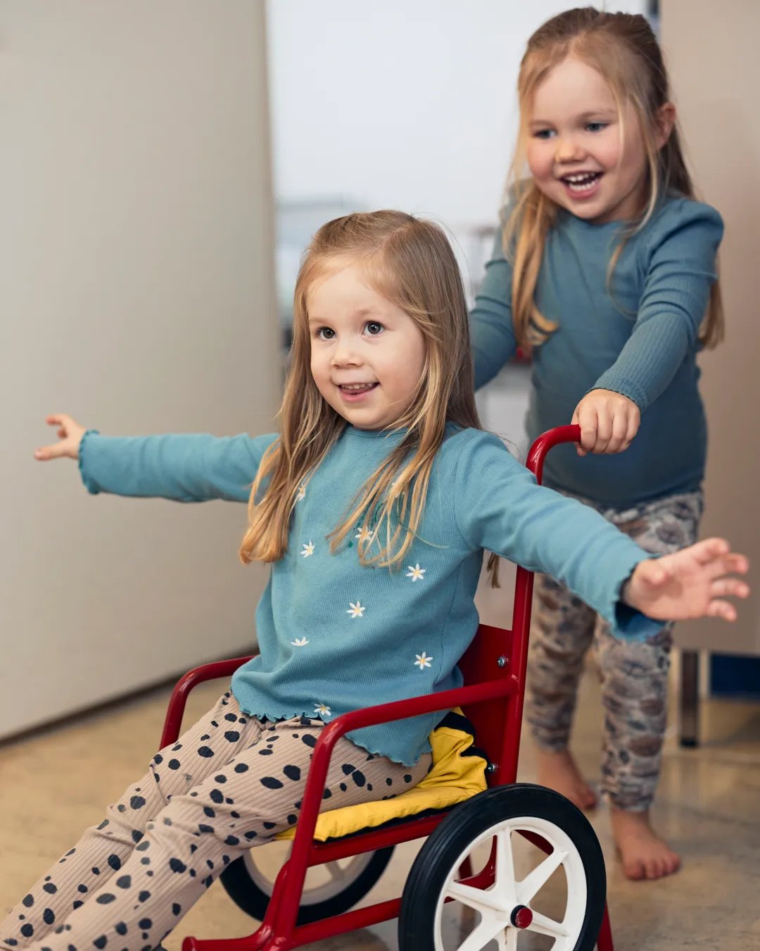 Två små flickor leker med en röd liten rullstol.