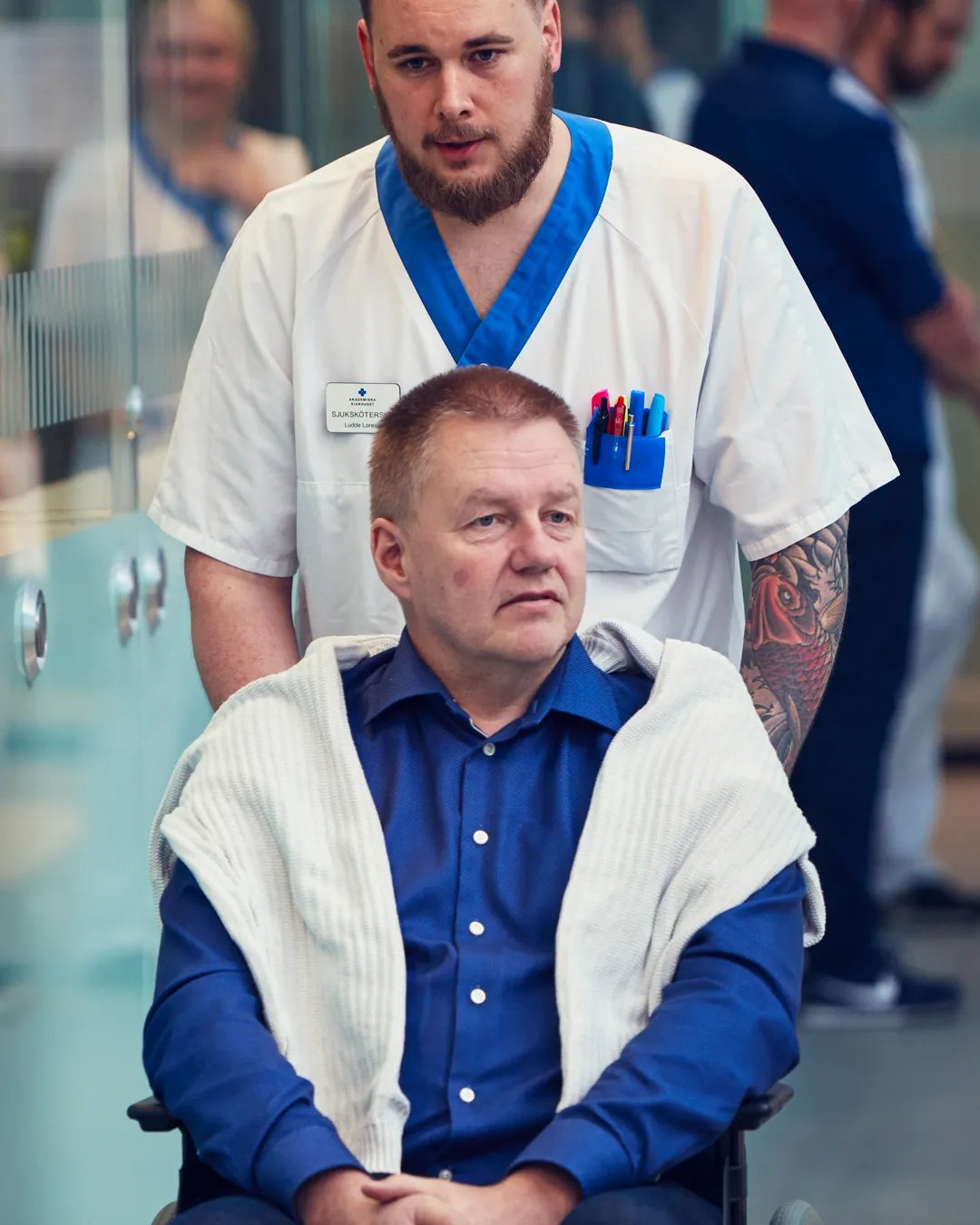 En patient blir skjutsad i rullstol av en man i vårdkläder.