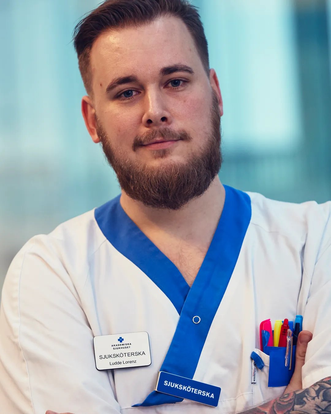 En manlig sjuksköterska med skägg och tatueringar som har vita arbetskläder.