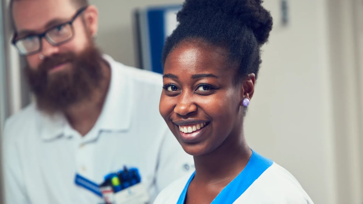 En kvinna i operationskläder ler medan en man i vit uniform tittar på.