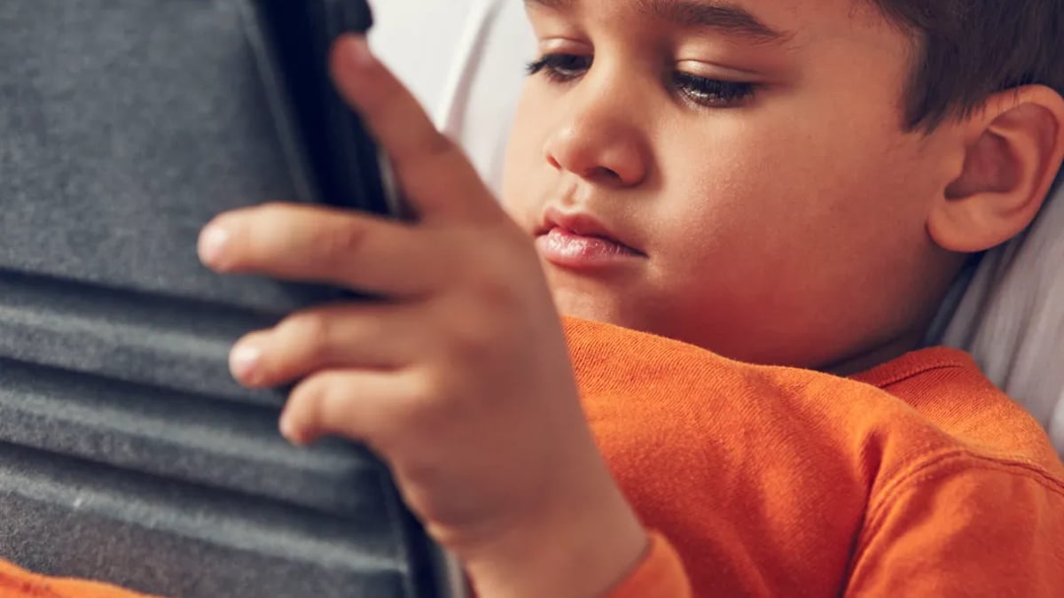 A young boy laying in bed looking at a tablet.