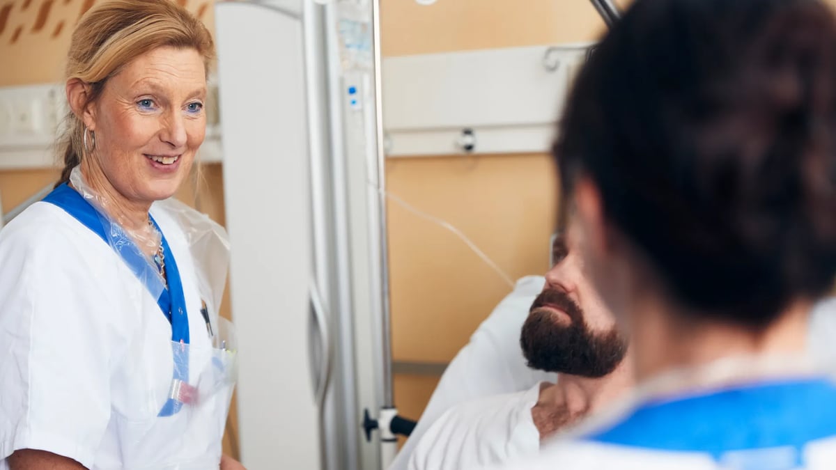 En kvinna i operationskläder pratar med en man i en sjukhussäng.