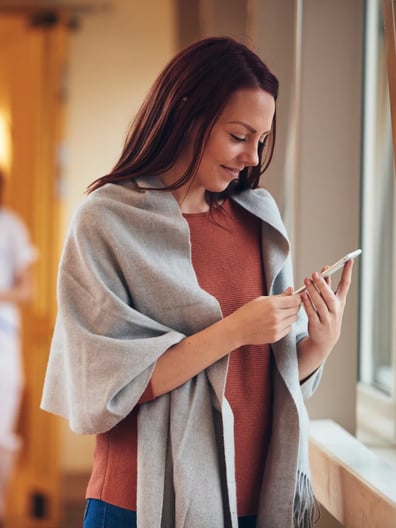 En kvinna står bredvid ett fönster och tittar på sin mobiltelefon.