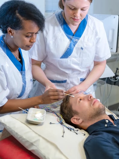 En patient ligger i en sjukhussäng och undersöks av två sjuksköterskor.