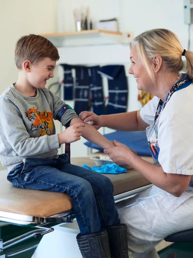 En sjuksköterska sitter på en stol bredvid en liten pojke som ler.