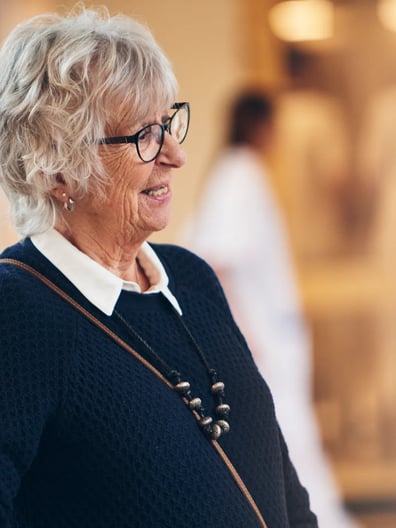 En äldre kvinna står i en sjukhuskorridor och ler.