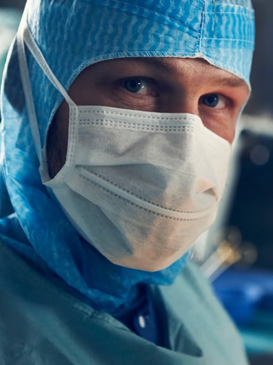 En kirurg har kirurgmask och operationskläder på sig och tittar in i kameran.