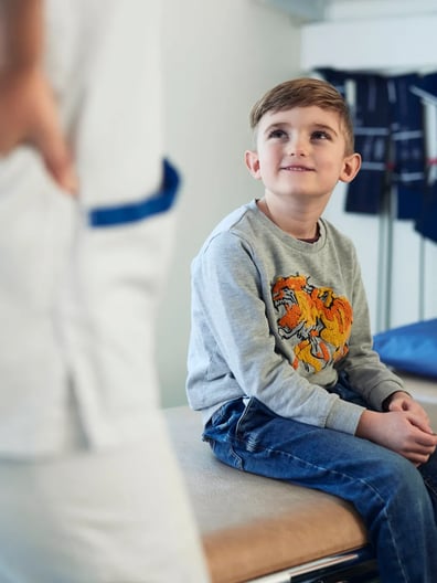 En ung pojke sitter på en sjukhussäng och tittar på en sjuksköterska med ett leende.