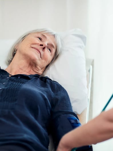 En äldre kvinna ligger i en sjukhussäng och ler mot en sjuksköterska.