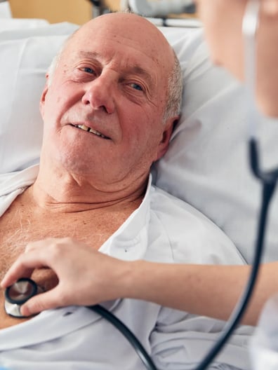 En man som ligger i en sjukhussäng blir undersökt med stetoskop.