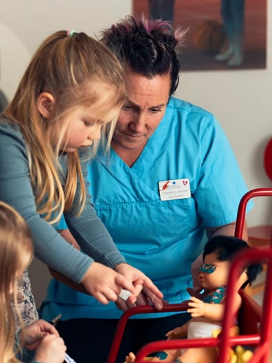 Två små flickor leker med en docka bredvid en vårdgivare.