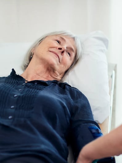 En äldre kvinna i en sjukhussäng tittar på en sjuksköterska med ett leende.