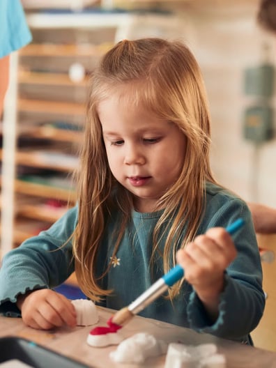 En liten flicka sitter vid ett bord med en pensel i handen och målar.
