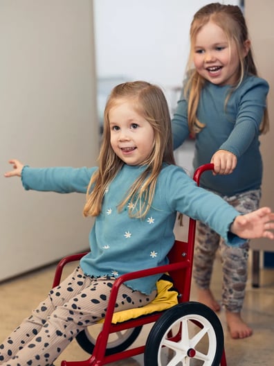 Två små flickor leker med en röd liten rullstol.