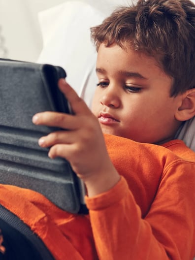A young boy laying in bed looking at a tablet.