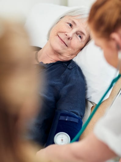 En äldre kvinna ligger i en sjukhussäng och ler mot kameran.