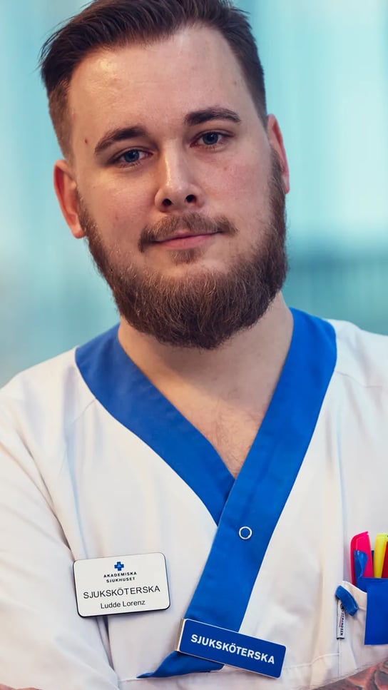 En manlig sjuksköterska med skägg och tatueringar som har vita arbetskläder.