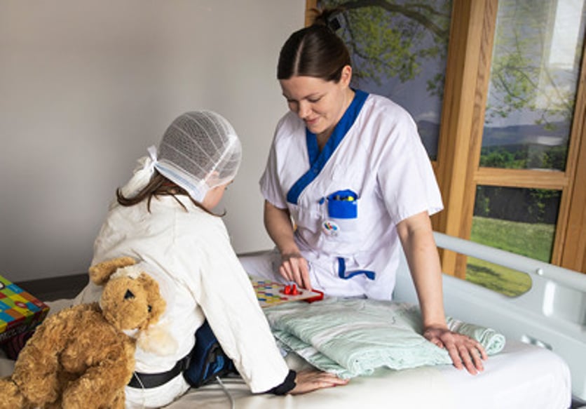 Ett barn har ett bandage om huvudet och sitter i en sjukhussäng och spelar spel med en sjuksköterska